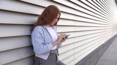 Canlı kızıl saçlı genç bir kadın, çarpıcı çizgili bir duvara yaslanarak modern, teknolojik bir yaşam tarzını somutlaştırarak ve sıradan bir hayat sürerek dikkatle akıllı telefonuna odaklanır.