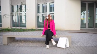 Canlı pembe ceketli şık bir genç kadın modern bir binanın dışındaki bankta şık bir kıyafet ve aksesuarla şık bir şekilde oturuyor ve kendine güveni tam.