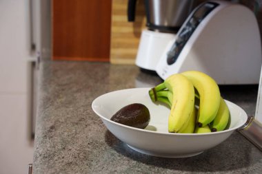 Taze meyve vitrinli mutfak, tezgahta muz ve avokado düzenleyen ev aşçısı, blender yakınındaki granit yüzeye yerleştirilmiş muz ve avokado.