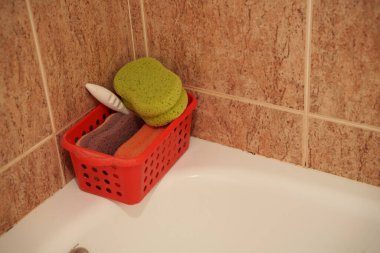 Küvetin köşesinde sünger sepeti renginde temizlik bezleri ve kırmızı plastik taşıyıcılarla bej kaplama duvara karşı yumuşak sıcak aydınlatma ev temizliği.