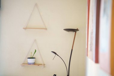 Minimalist, rahat bir kitap köşesi, bitkilerin sergilendiği üçgen bir raf, uzun siyah zemin lambasının yanında dokulu nötr duvara karşı sulu bir saksı tutuyor.,