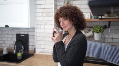 Güzel kıvırcık saçlı düşünceli bir kadın şık ve modern mutfağında duruyor, elinde bir kamera ve yansıtıcı bir şekilde bakıyor, yaratıcılık ve ilham dolu sakin bir anı yakalıyor.