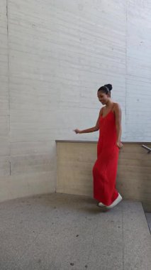 Dalgalı kırmızı elbiseli neşeli genç bir kadın modern moda ve yaratıcılığı harmanlayan şehir ortamında neşeli bir poz veriyor. Neşeli tavırları mutluluk saçıyor ve güven veriyor.