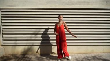 Neşeli ve kendinden emin genç bir kadın canlı ve göz kamaştırıcı kırmızı bir elbise içinde modern dokulu bir duvara karşı zarif bir şekilde poz vererek pozitiflik, tarz ve neşe saçıyor.