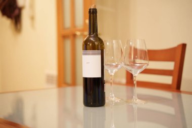 Şarap şişesi masada iki bardak, sıcak mutfak ışığı, arka planda ahşap sandalye, boş etiket, sığ alan derinliği, akşam yemeği öncesi ayarları samimi olduğunu düşündüren