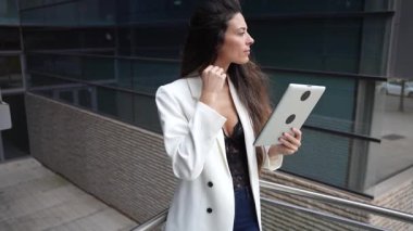 In a vibrant urban setting, a stylish woman in a white blazer is deeply focused on her tablet, reflecting her professionalism and the integration of technology into her modern lifestyle