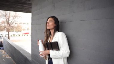 Genç bir kadın, bir elinde şık bir kahve fincanı diğer elinde bir tablet, çağdaş kentsel tarzı ve profesyonelliği somutlaştıran modern gri bir duvara karşı durmaktadır.