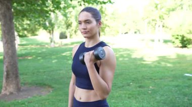 Genç, formda bir kadın, güneşli bir parkta, kas gücü ve formda bir halterle biseps yapıyor. Kararlılık saçıyor ve aktif açık hava egzersizlerine odaklanıyor.