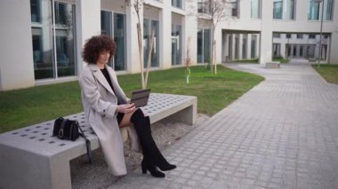 Manzaralı bir açık hava ortamında, şık genç bir kadın dizüstü bilgisayarına dalmış bir şekilde bankta oturuyor, güzellik ve modern teknolojinin günlük yaşam üzerindeki etkisini gözler önüne seriyor.