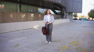 Moda kıyafetleri içinde genç bir kadın renkli sonbahar yapraklarıyla kaplı yoğun bir kaldırımda güven içinde duruyor, modern mimari hareketli şehir ortamının huzurlu atmosferini yükseltiyor.