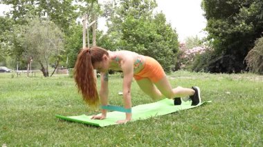 Bir kadın, yemyeşil bir parkla çevrili yoga minderindeki bir direniş bandını kullanarak dışarıda antreman yapıyor. Fitness, sağlık ve egzersiz temaları için mükemmel
