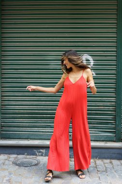 Şık bir kadın dikkat çekici derecede koyu kırmızı bir tulumla poz veriyor, canlı yeşil bir arka plana karşı çağdaş şehir modasını sergiliyor, inanılmaz göz alıcı bir görsel etki yaratıyor.