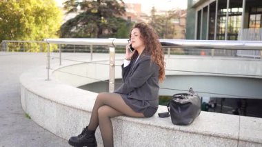 Modern mimari ve yeşillik ile çevrili şık bir iş kadını telefonda konuşuyor. Diğerlerine ilham veren, güven ve başarıyı somutlaştıran canlı bir şehir havası yaratıyor.