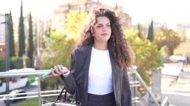 Güzel kıvırcık saçlı dengeli ve kendine güvenen bir iş kadını şehir manzarasına karşı gururla duruyor. Modern moda anlayışını ve profesyonelliğini yansıtan kendine özgü tarzını sergiliyor.