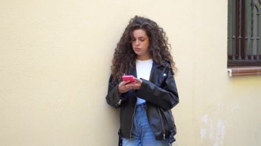 Şık deri ceketli ve şık şortlu şık kıvırcık saçlı genç bir kadın parlak sarı bir duvara yaslanmış, akıllı telefonuna odaklanmış, modern yaşam tarzını ve modasını somutlaştırıyor.