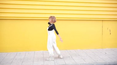 Modayı takip eden beyaz tulumlu neşeli ve hayat dolu bir insan şık bir siyah bluzla uyumlu, neşeli ve eğlenceli bir pozu canlı sarı bir duvara yapıştırıyor. Bu da sanatsal etkiyi arttırıyor.