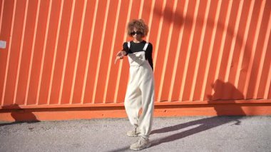 Eşsiz bir stil sahibi, modaya uygun tulumlar giyen, parlak turuncu bir duvara karşı neşeli bir duruş sergiliyor ve büyüleyici ve dinamik bir görsel başyapıt ortaya çıkıyor.