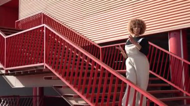 Şık bir şahıs canlı kırmızı bir merdivende zarif bir şekilde poz verir, yüksek moda ve sanatsal yaratıcılığın şaşırtıcı bir karışımını hayat ve renklerle dolu canlı bir şehir ortamında sergiler.