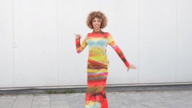 Canlı kıvırcık saçlı neşeli bir kadın neşeli bir arka plana karşı renkli bir elbise içinde poz verir, ruhunu ve moda anlayışını sergiler, görüntüyü büyüleyici ve hayat dolu yapar.