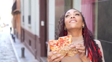 Şık bir kadın canlı şehir caddesinde durmuş büyük, lezzetli bir dilim pizzaya hayran. Renkli ve canlı saçları onu çevreleyen enerjik atmosferi mükemmel bir şekilde güçlendiriyor.