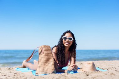 Güneşli bir günde, şık güneş gözlüklü genç bir kadın renkli bir plaj havlusunda dinlenir, güneşi emer ve dalgaları dinler, bu huzurlu ortamda neşe ve rahatlığı somutlaştırır.