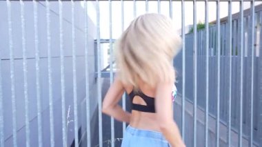 Şık bir kot ve renkli bir bluz giyen mutlu, genç bir sarışın kadın güneşli bir günde hareketli bir şehirde canlı kırmızı pencereleri olan çağdaş bir binanın yanında yürüyor.