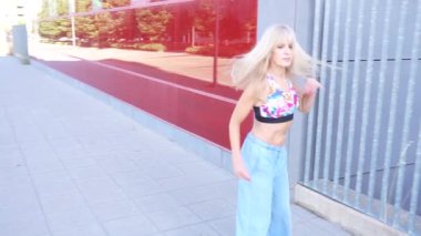 Şık, sarışın bir kadın zarif bir şekilde modern bir şehir binasının yanında yürüyor. Şık ve kendinden emin bir hava yayıyor. Şehir ortamında, çağdaş bir moda ve sakin bir şehir yaşam tarzı sergiliyor.