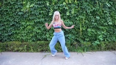 Canlı bir açık hava sahnesinde dinamik bir kadın enerjik bir şekilde kot pantolon ve renkli bir hasat bluzu ile sarmaşık kaplı bir duvara karşı dans eder. Kaygısız ve neşeli hareketleriyle neşe ve coşku saçar.