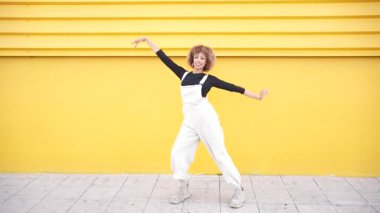 Güzel kıvırcık saçlı, zarif, genç bir kadın, kendine güvenen bir duruş sergiliyor. Canlı ve neşeli sarı bir duvarın önünde. İnanılmaz eşsiz stili ve hayat dolu kişiliğini sergiliyor.