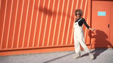 Harika bir stil anlayışı olan bir kişi şık siyah bir üst ile şık bir tulum içinde, modern, güncel bir havayı ortaya çıkaran canlı turuncu bir duvara karşı durarak rahatça poz verir.