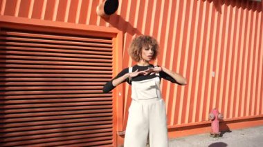 Moda düşkünü genç bir kadın kendine güvenerek ve gururla renkli turuncu bir duvarın önünde duruyor. Dünyaya özel stilini, modern estetiğini ve canlı kişiliğini sergiliyor.