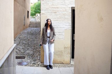 Sokakta bavulu olan genç bir kadın, sıradan bir gezgin, taş duvara karşı duruyor telefon ve bavullar, gevşemiş güneş ışığı havası, dar kaldırım, dokulu.