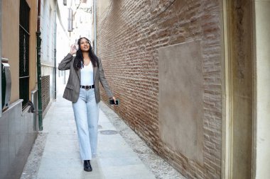 Tuğla duvarda yürüyen bir kadın telefonu tutuyor, dar şehir geçidinde kasıtlı tempo, tarafsız spor ceket ve kot, odaklanmış ifade, samimi banliyö