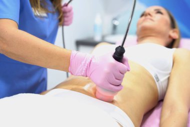 Estetisyen ile ultrason karın tedavisi gören kadın jel ve el bilgisayarı, pembe eldivenler, tedavi yatağında yakın çekim göbeği, modern klinik.