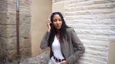 Şık bir kadın en sevdiği kulaklıklarıyla canlı şehir sahnesinden hoşlanır. Zarif bir şekilde şık bir ceket ve çarpıcı bir beyaz kıyafet giyer ve büyüleyici bir sokakta dokulu bir duvara karşı durur.