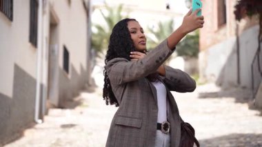 Şık giyinmiş, kendine güvenen bir kadın, bir sokak arasında yürüyor, elinde bir akıllı telefon tutuyor, güçlü bir özgüven duygusu yayıyor ve yürürken dengede duruyor.