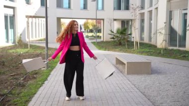 Modaya uygun pembe ceketli ve parlak siyah pantolonlu şık bir kadın modern bir avluda duruyor. Yeşillik, kendine güven ve cazibe saçıyor. Zarafetiyle seyircileri büyülüyor.