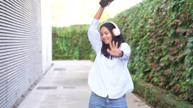 Neşeli bir kadın, gündelik giyinmiş ve kulaklık takmış, canlı, yemyeşil bir arka planla çevrili iken müziğe dalmış, güzel ve kaygısız bir keyif ortamı yaratıyor.