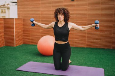 Diz çökmüş bir kadın damdaki dambılları kaldırıyor, omuzlarını kaldırıyor, odaklanmış ifade, siyah hasat bluzu ve tozluklar, turuncu denge topu.