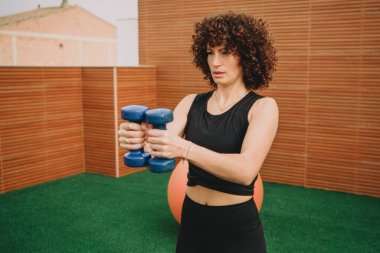 Göğüs kafesinde dambıl tutan kadın antrenör, odaklanmış ifade, denge topunun yanında yeşil çimlerde duran tahta paneller, düşük etki gücü rutini,