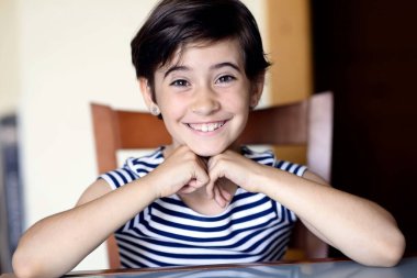 Kahvaltı masasında ışıldayan çocuk, elleri katlanmış ve gülümsüyor, çizgili gömlek ve parlak gözler, yüzünde sabah güneşi, ahşap sandalye ve mutfak arka planı, samimi