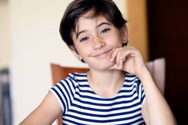 Masada gülümseyen bir çocuk, düşünceli bakışlar, çizgili gömlek, eline yaslanma, sıcak doğal ışık, rahat mutfak arka planı, ince bir bokeh, sakin bir sabah havası, odaklanmış.