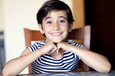 Masada sırıtan çocuk, elleri çenenin altına sıkışmış, parlak gözler ve açık ifade, çizgili gömlek ve rahat duruş, sıcak iç aydınlatma, davetkar bir ortam