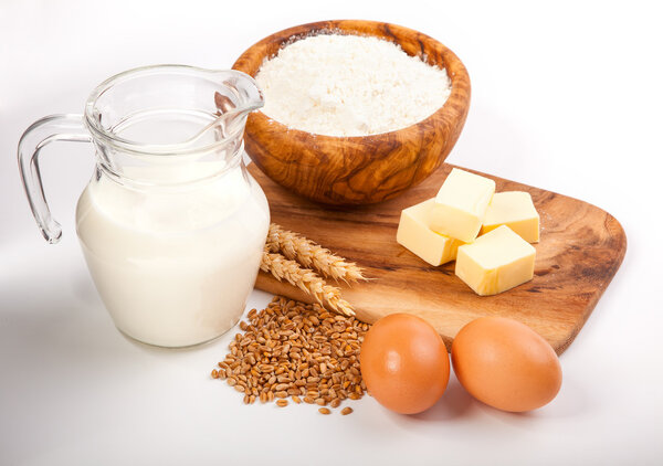 Glass jug with milk, wheat seeds, flour and two eggs on white ba