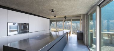 Büyük adası olan modern beyaz mutfak, indüksiyon hob, iki fırın, lavabo ve paslanmaz çelik kapak. İsviçre 'nin doğasına bakan büyük pencereler. İçeride kimse yok.