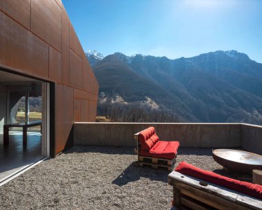 Verandası olan modern bir ev. İsviçre dağlarına bakan kırmızı kanepe. İçerde kimse yok.