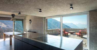 Büyük adalı modern beyaz mutfak, indüksiyon hob, lavabo ve paslanmaz çelik kapak. İsviçre 'nin doğasına bakan büyük pencereler. İçeride kimse yok.