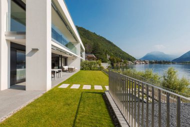 Ceresio Gölü 'nün hemen yanında bahçeli iki dairesi olan modern bir ev. Mavi gökyüzü güneşli. Minimalist ve doğrusal mimari. Gölün panoramik manzarası. Metnin için boşluk
