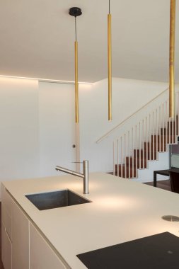 Modern tasarım mutfağının ayrıntıları, siyah indüksiyon pişirme, paslanmaz çeliğe batma ve tasarım lambası. Arka planda kapalı bir kapı ve kantilever merdiven var. Beyaz duvar. İçeride kimse yok.