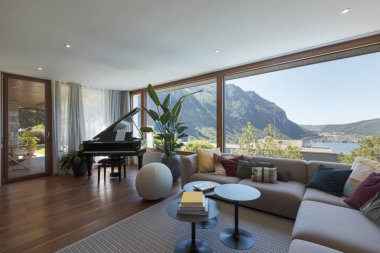 Oturma odasında büyük açık renkli bir kanepe, bir sürü yastık ve siyah bir piyano var. Göl manzaralı vadiye bakan büyük bir pencere. İçeride kimse yok. 